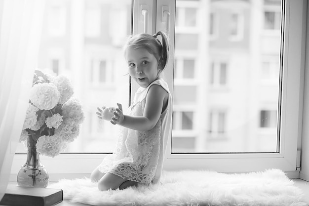 Uma garotinha está sentada no parapeito da janela. Um buquê de flores em um vaso perto da janela e uma garota cheirando flores. Uma princesinha em um vestido branco com um buquê de flores brancas na janela.