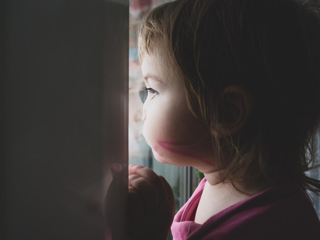 Foto uma garotinha está triste e olha pela janela, ela quer andar na rua. a criança olha pela janela. retrato de um bebê triste no parapeito da janela.