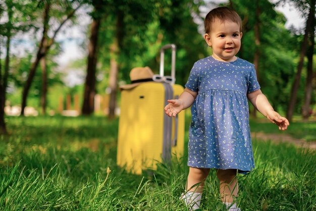 Uma garotinha feliz em um vestido azul de verão fica ao lado de uma mala e plantas verdes