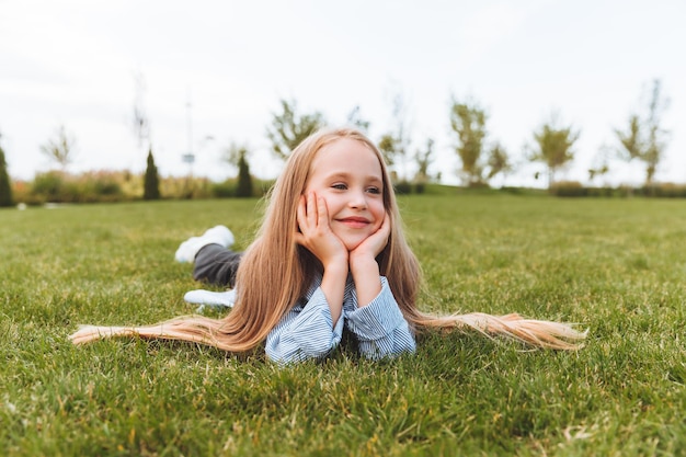 Uma garotinha feliz em uma jaqueta jeans está no gramado verde uma criança sorridente está descansando deitada na grama o conceito de uma infância despreocupada