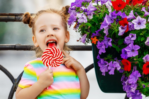 Uma garotinha fica perto de um canteiro de flores e lambe um pirulito na mão