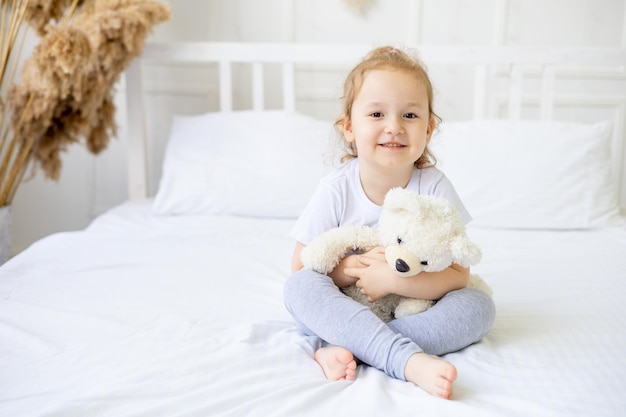 Uma garotinha fofa em uma cama de algodão branco em casa com um brinquedo macio nas mãos sorrindo na cama em casa