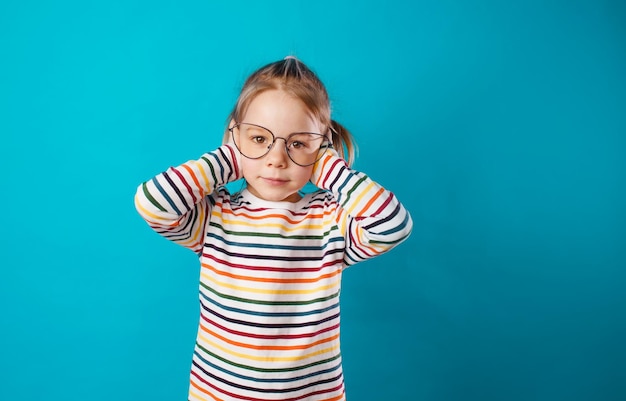 Uma garotinha linda de óculos grandes cobre os ouvidos com as mãos em um fundo azul isolado