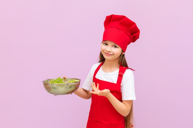 Uma garotinha linda fantasiada de chef vermelho está comendo uma salada fresca A criança adora cozinhar em casa