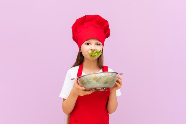 Uma garotinha linda fantasiada de chef vermelho está comendo uma salada fresca A criança adora cozinhar em casa