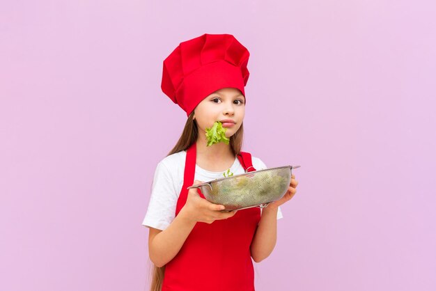 Uma garotinha linda fantasiada de chef vermelho está comendo uma salada fresca A criança adora cozinhar em casa