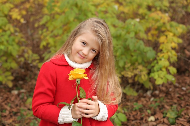 Uma garotinha loira com um casaco vermelho segura uma rosa amarela nas mãos no parque