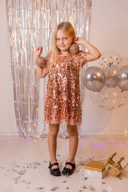 uma garotinha loira com um vestido brilhante brinca com bolas de natal no carnaval de ano novo