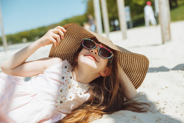 Uma garotinha loira de chapéu de palha está em uma praia de areia tomando sol na areia