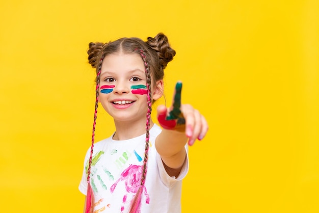 Uma garotinha manchada em tintas multicoloridas para desenhar estica o dedo indicador para a frente apontando para o seu recalama em um fundo amarelo isolado O desenvolvimento da arte e da criatividade infantil