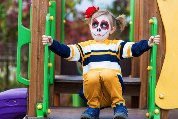 Uma garotinha pré-escolar com o rosto pintado, mostra caretas engraçadas no playground, comemora o halloween ou o dia mexicano dos mortos.