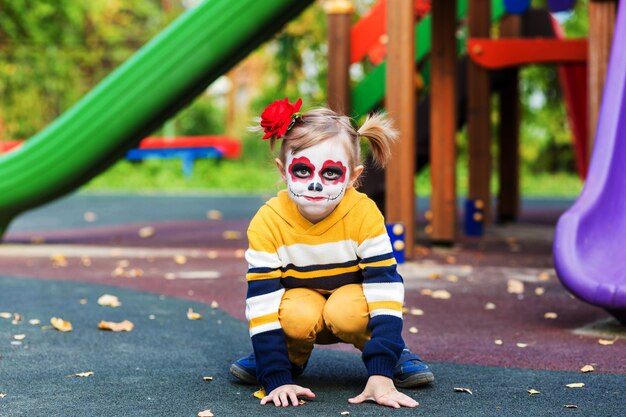 Uma garotinha pré-escolar com o rosto pintado, sorrindo no parquinho, comemora o Halloween ou o dia mexicano dos mortos.