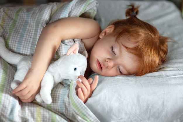 Uma garotinha ruiva dorme pacificamente à noite em sua cama e abraça um gatinho de pelúcia