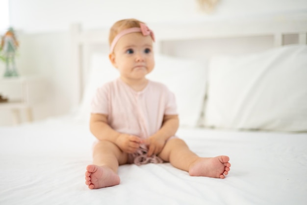 Uma garotinha saudável bonitinha de até um ano de idade em uma roupa rosa feita de tecido natural está sentada em uma cama na roupa de cama branca no quarto olhando para a câmera o bebê está em casa