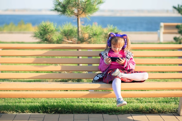 Uma garotinha se senta em um banco do parque e olha para os desenhos do telefone através de fones de ouvido sem fio. O problema das crianças modernas com gadgets