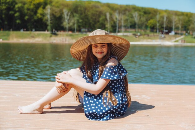 Uma garotinha se senta em um píer perto do lago da cidade ao ar livre nas férias de verão