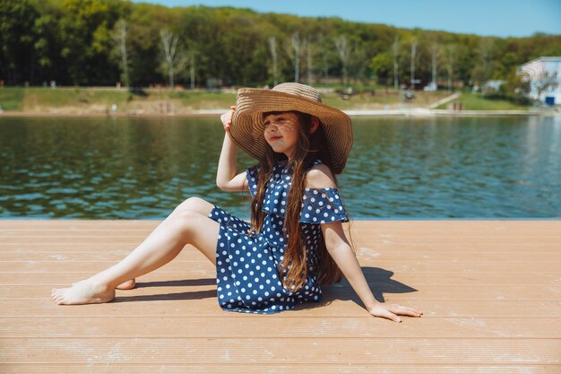 Uma garotinha se senta em um píer perto do lago da cidade ao ar livre nas férias de verão