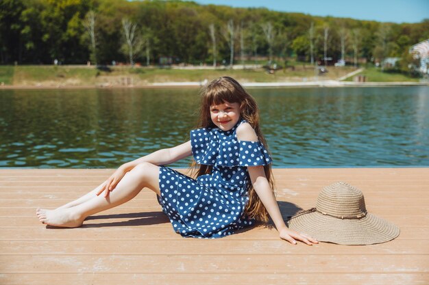 Uma garotinha se senta em um píer perto do lago da cidade ao ar livre nas férias de verão