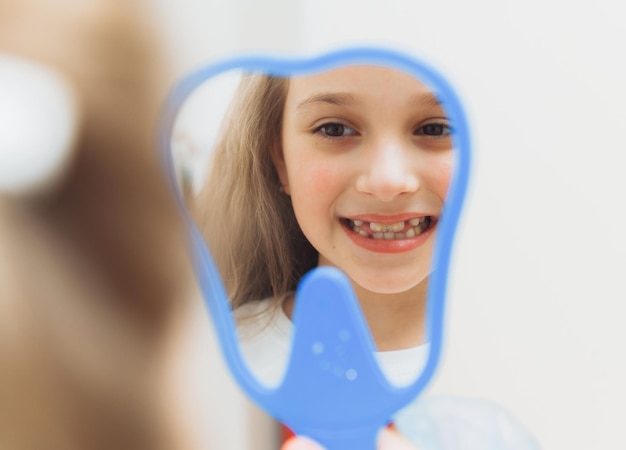 Uma garotinha se senta em uma cadeira odontológica e examina seus dentes curados enquanto olha no espelho
