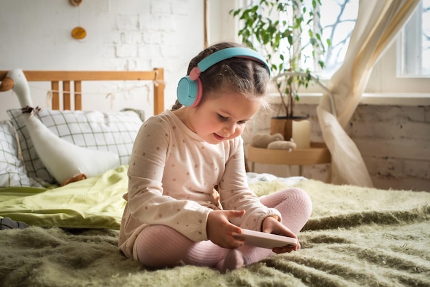 Uma garotinha se senta na cama em fones de ouvido bebê imprudentemente joga jogos e ouve música