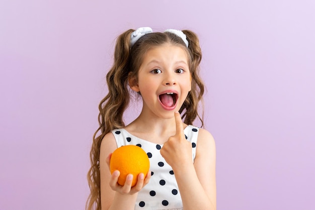 Uma garotinha segura laranjas nas mãos e aponta para a boca em um fundo rosa isolado Frutas cítricas para comida de bebê