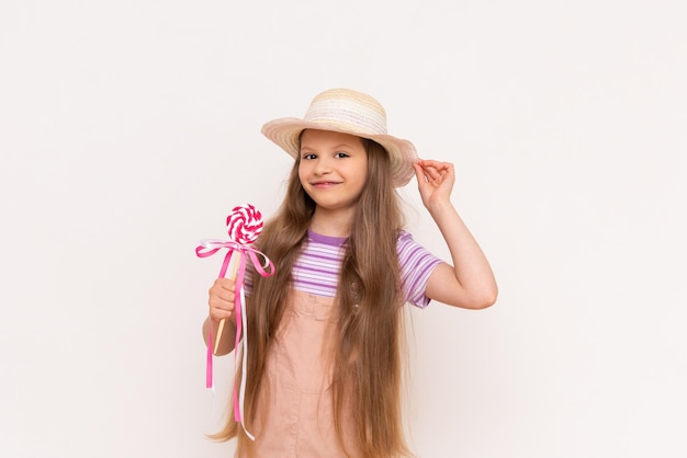 Uma garotinha segura um pirulito de caramelo em uma vara no bebê em um vestido de verão e um chapéu de palha de verão em um fundo branco isolado