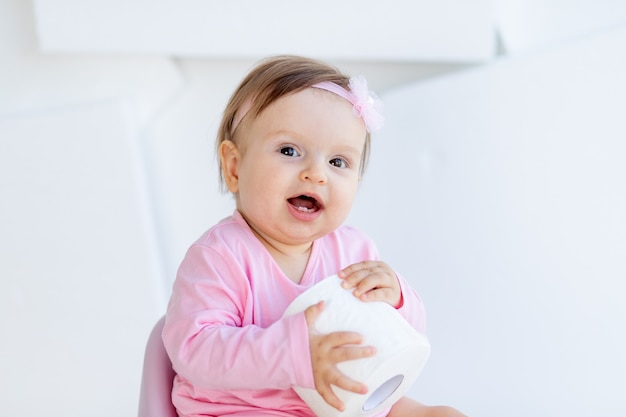 Uma garotinha sentada em um penico com roupas cor de rosa em uma sala iluminada e brincando com papel higiênico