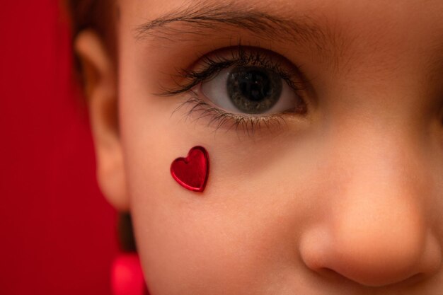 Uma garotinha séria olha para a câmera com o coração na bochecha com tranças em um fundo vermelho Foto de alta qualidade