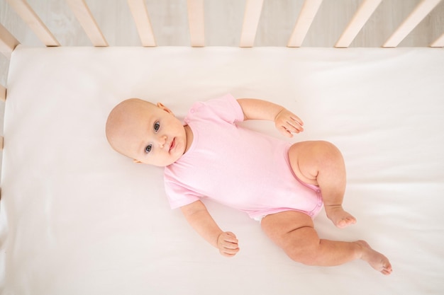 Uma garotinha sorridente e fofa em uma roupa rosa está deitada em um berço em casa de costas olhando para a câmera sorrindo bebê feliz