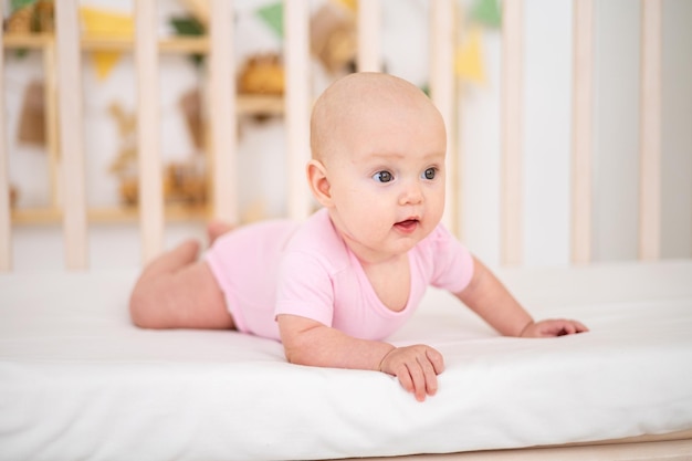 Uma garotinha sorridente e fofa em uma roupa rosa está deitada em um berço em casa na cama branca de bruços olhando para a câmera sorrindo bebê feliz