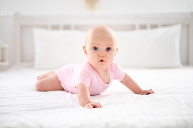 Uma garotinha sorridente e fofa em uma roupa rosa está deitada na cama no quarto da casa na cama branca de bruços olhando para a câmera sorrindo bebê feliz