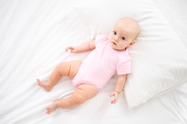 Uma garotinha sorridente e fofa em uma roupa rosa está deitada na cama no quarto da casa na cama branca de costas olhando para a câmera sorrindo bebê feliz