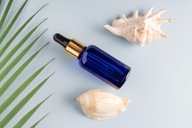 Uma garrafa de vidro com óleo facial ou soro em um fundo azul com conchas e folha de palmeira tropical Conceito cosmético de spa orgânico natural Vista superior