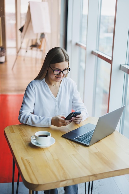 Uma gerente de mulher inteligente com uma xícara de café tem uma conversa de negócios por meio de um smartphone para discutir um pedido de entrega uma jovem proprietária se senta em uma mesa em um café com um laptop e liga