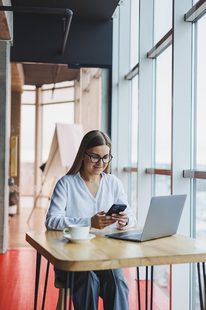 Uma gerente de mulher inteligente com uma xícara de café tem uma conversa de negócios por meio de um smartphone para discutir um pedido de entrega uma jovem proprietária se senta em uma mesa em um café com um laptop e liga