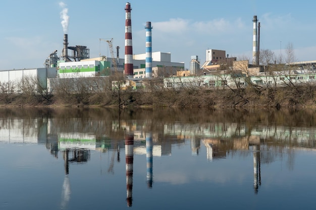 Uma grande fábrica está localizada na margem do rio Fumaça branca tóxica escapa das chaminés da fábrica O risco de um desastre ambiental Reflexo na água do rio da fábrica