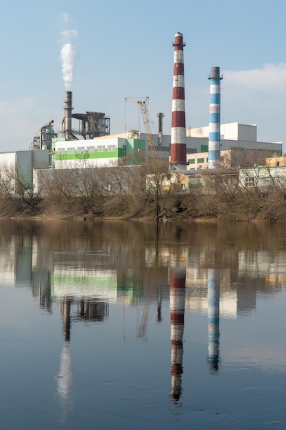 Uma grande fábrica está localizada na margem do rio Fumaça branca tóxica escapa das chaminés da fábrica O risco de um desastre ambiental Reflexo na água do rio da fábrica