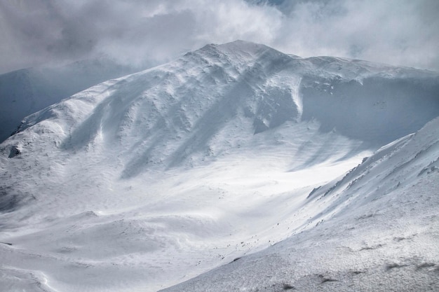 Uma grande montanha rochosa branca com um couloir escuro está coberta de neve entre as nuvens nebulosas antes da nevasca de inverno