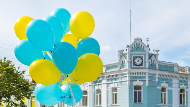 Uma grande pilha de balões de hélio amarelo e azul contra o céu e uma casa com um relógio ao fundo