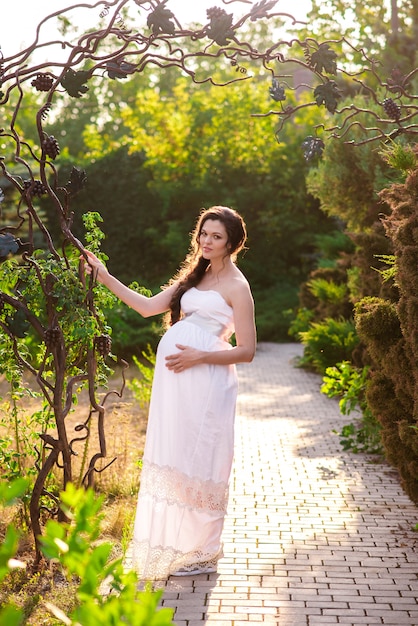 Uma grávida vestida de branco abraça a barriga e sorri no parque, o sol brilha e a grama verde. Conceito de uma mulher feliz e de maternidade.