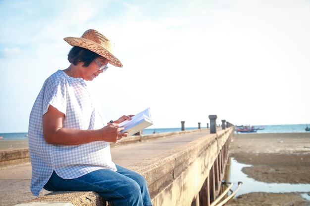 Uma idosa mulher asiática usando um chapéu para ler um livro Sentada à beira-mar, bom tempo, refrescante, boas festas