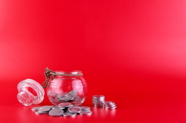 Uma jarra com moedas dentro e muitas moedas do lado de fora da jarra isoladas em um fundo vermelho