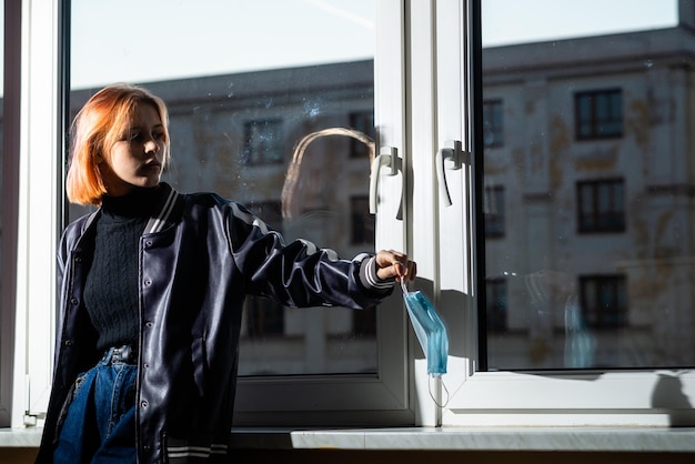 Uma jovem adolescente com uma máscara de medina senta-se no parapeito da janela Esperando solidão depressão tédio Anos adolescentes