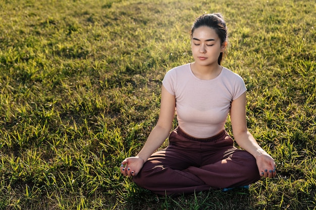 Uma jovem adulta de aparência asiática senta-se no gramado e medita.