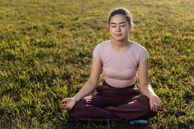 Uma jovem adulta de aparência asiática senta-se no gramado e medita.