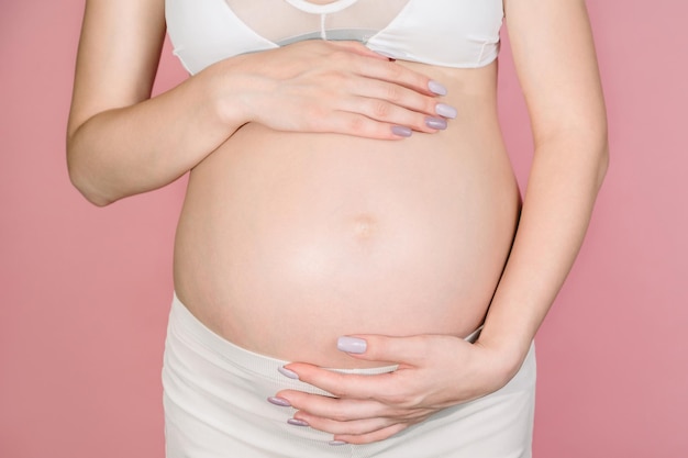 Uma jovem adulta grávida em roupas brancas toca sua grande barriga nua com as mãos, mostrando a forma o conceito de gravidez closeup isolado em um fundo rosa