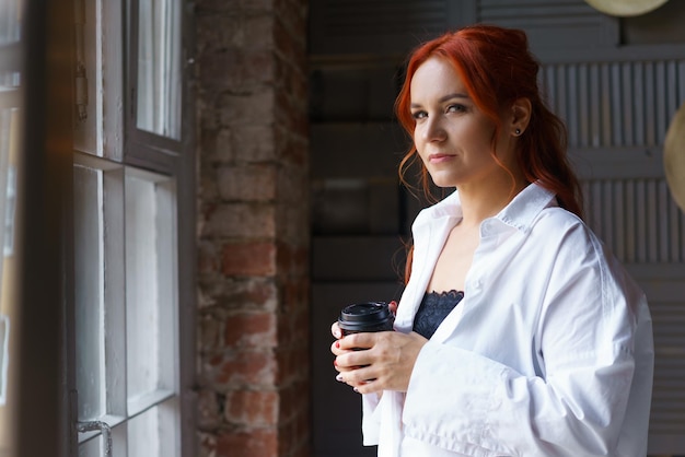 Uma jovem adulta ruiva com uma camisa branca em pé na janela e segurando um copo de café