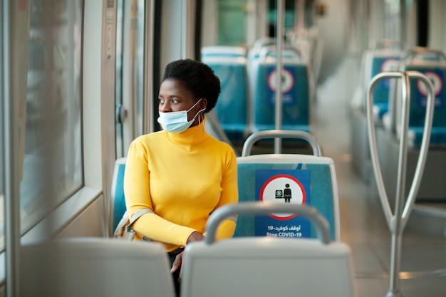 Uma jovem africana usando uma máscara protetora viaja em um ônibus e olha pela janela. Coronavírus, distância social.