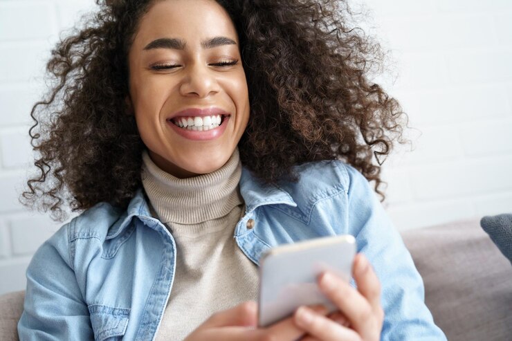 Foto uma jovem afro-americana alegre segura um telefone inteligente usando chamadas de vídeo do aplicativo assistindo vídeos engraçados nas mídias sociais feliz jovem adolescente africana jogando jogo para celular rindo olhando no celular em casa