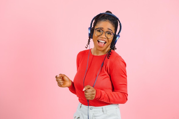 Uma jovem afro-americana com um sorriso brilhante vestida com óculos de roupas casuais e fones de ouvido dança sobre fundo rosa